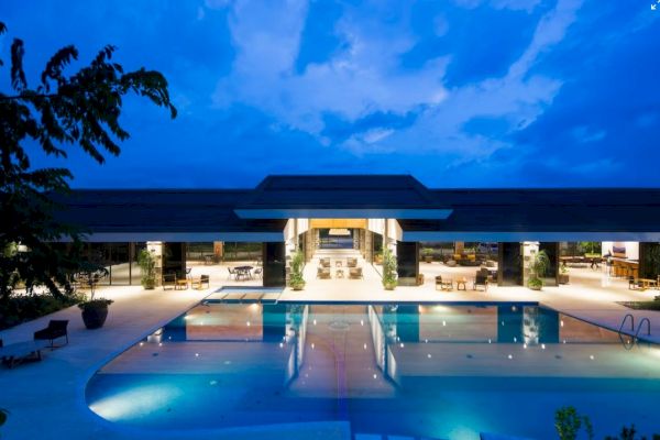 A luxurious resort with a large, illuminated swimming pool, lounge chairs, and a spacious, open-air building at dusk.