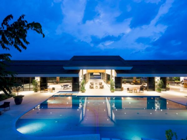 A luxurious resort with a large, illuminated swimming pool, lounge chairs, and a spacious, open-air building at dusk.