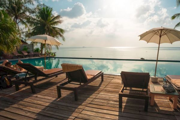 A tropical resort with sun loungers and parasols by an infinity pool overlooking the ocean. Palm trees and a beautiful sky complete the scene.