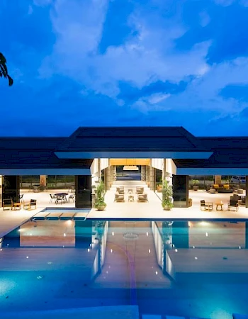 The image shows a luxurious resort with a large swimming pool, outdoor seating, and a modern building illuminated in the evening.