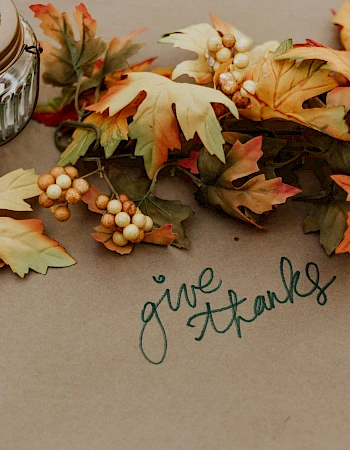 Autumn leaves and berries surround a small jar with "give thanks" written underneath in green script, creating a Thanksgiving-themed scene.