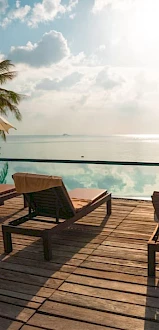 A serene poolside setting with lounge chairs, umbrellas, and a view of the ocean under a partly cloudy sky, surrounded by palm trees.