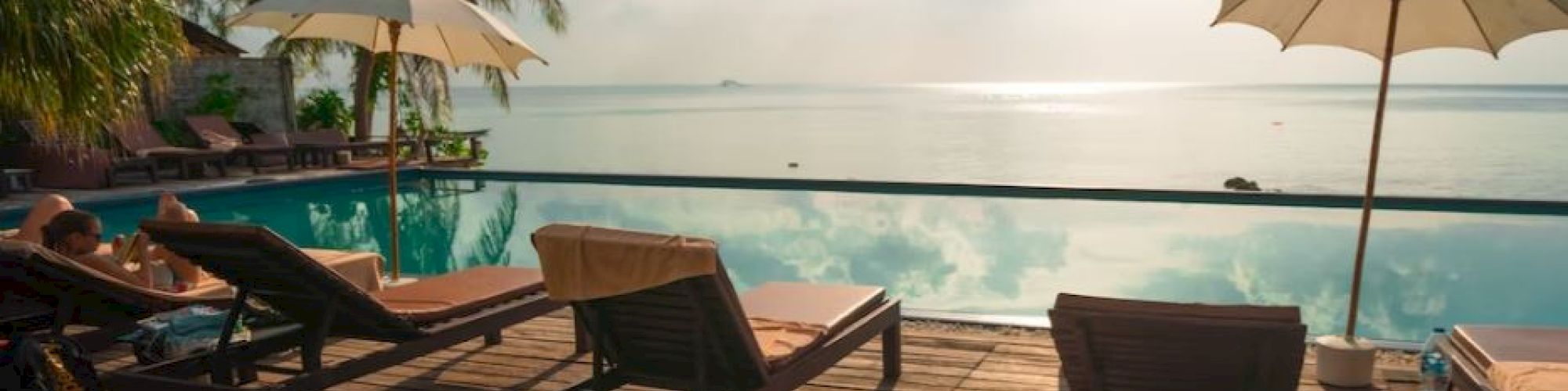 A serene poolside scene with lounge chairs, umbrellas, palm trees and a scenic view of a calm body of water at sunrise or sunset.