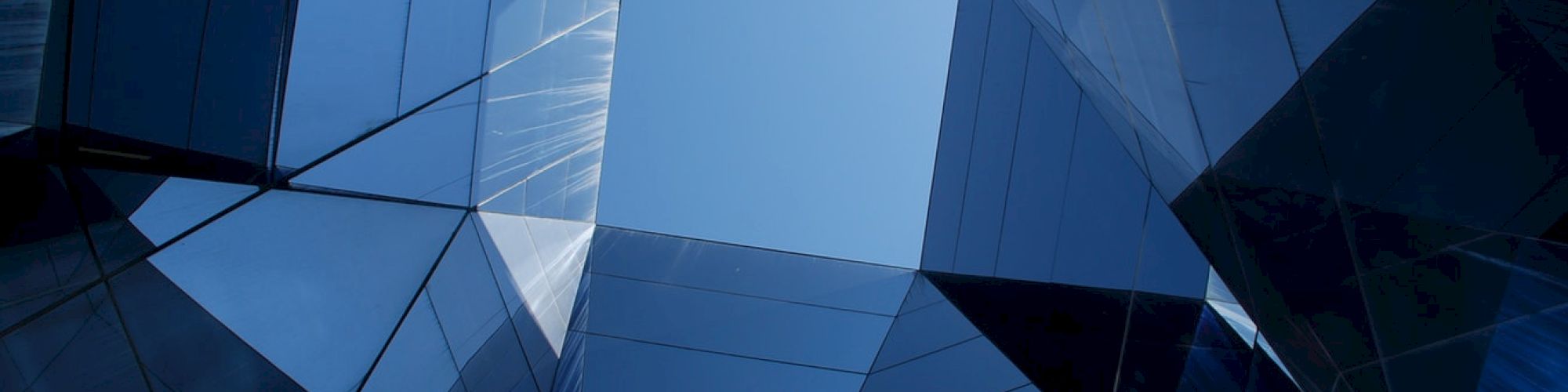 The image shows a view of the sky through tall, reflective glass buildings forming geometric patterns.