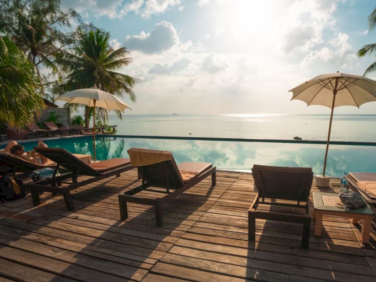 A serene poolside scene with lounge chairs, umbrellas, and a wooden deck facing a calm body of water under a partly cloudy sky.