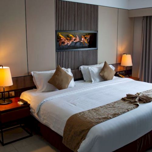 A modern hotel room with a large bed, decorative pillows, two bedside tables with lamps, and a lit wall feature above the bed.