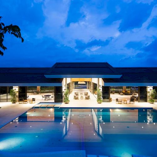 An illuminated luxury villa with a large swimming pool and outdoor seating area at dusk, featuring modern architecture and serene surroundings.