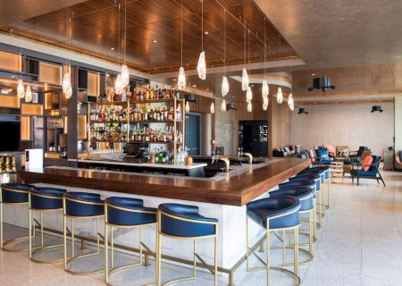 A modern bar with wooden counters, blue chairs, hanging lights, and various bottles on shelves, with a seating area in the background.