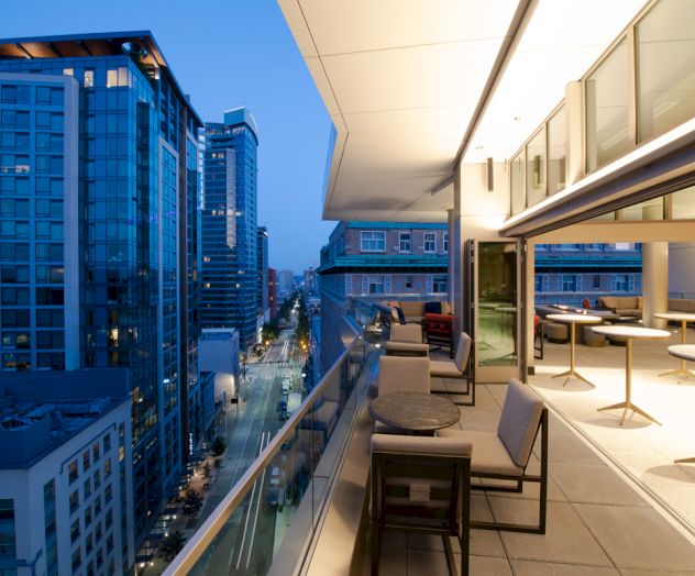 An outdoor terrace at dusk with tables and chairs, overlooking a city street lined with modern buildings. The scene is well-lit and inviting.