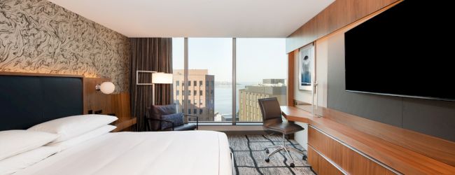 The image shows a modern hotel room with a large bed, a wall-mounted TV, a desk with a chair, and a view of buildings through large windows.