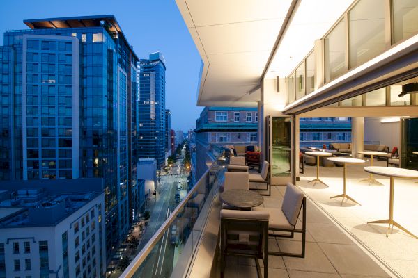 A cityscape view from a modern balcony at dusk, showing tall buildings, a street below, and an elegant outdoor seating area.