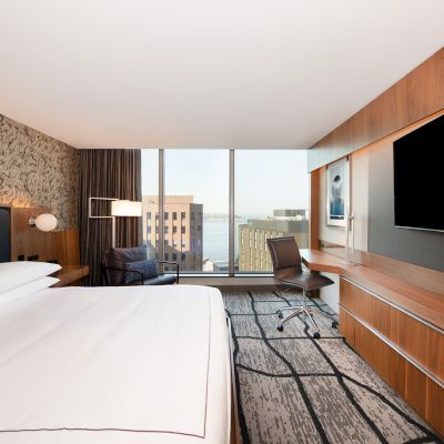 A modern hotel room with a large bed, wall-mounted TV, desk, and chair, featuring large windows with a cityscape view and a contemporary design.