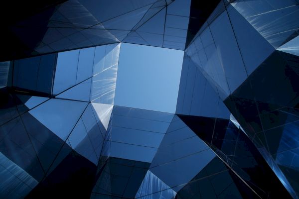 The image shows an upward view of modern glass buildings reflecting the sky, creating geometric patterns with sharp angles and blue hues.