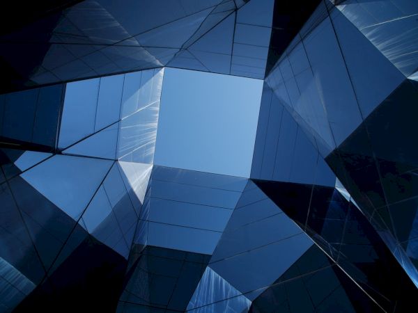 The image shows an upward view of modern glass buildings reflecting the sky, creating geometric patterns with sharp angles and blue hues.