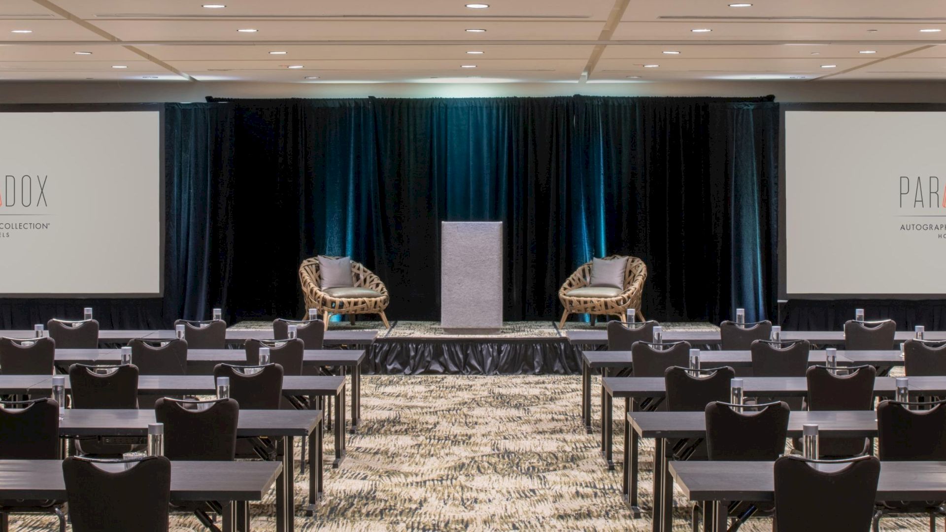 A conference room with rows of tables and chairs facing a stage with two chairs, a podium, and two screens displaying "PARADOX."
