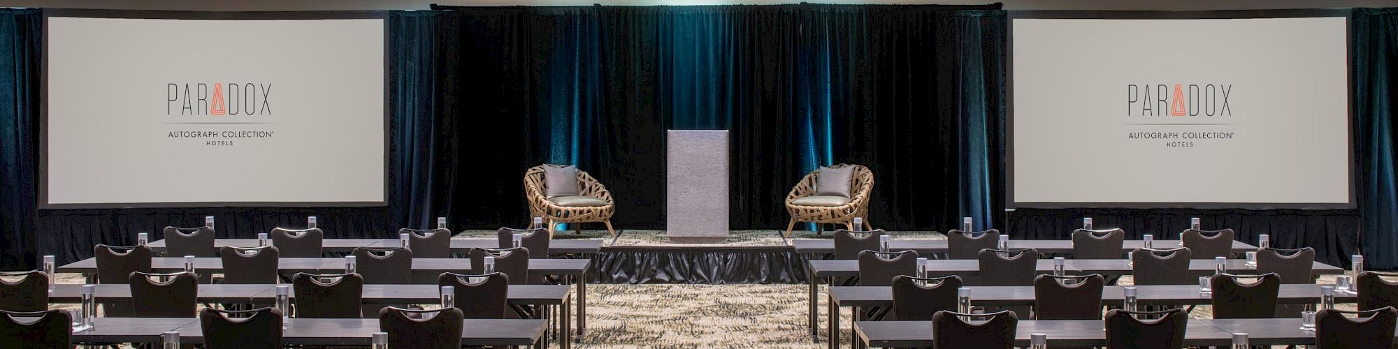 A conference room with rows of tables and chairs facing a stage with two chairs, a podium, and two screens displaying 