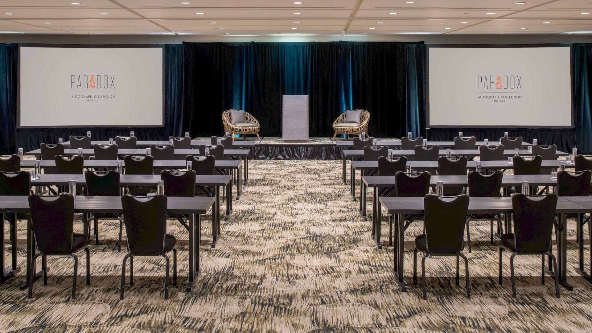 A conference room setup with rows of tables and chairs facing a stage with two screens labeled "PARADOX" and two armchairs.