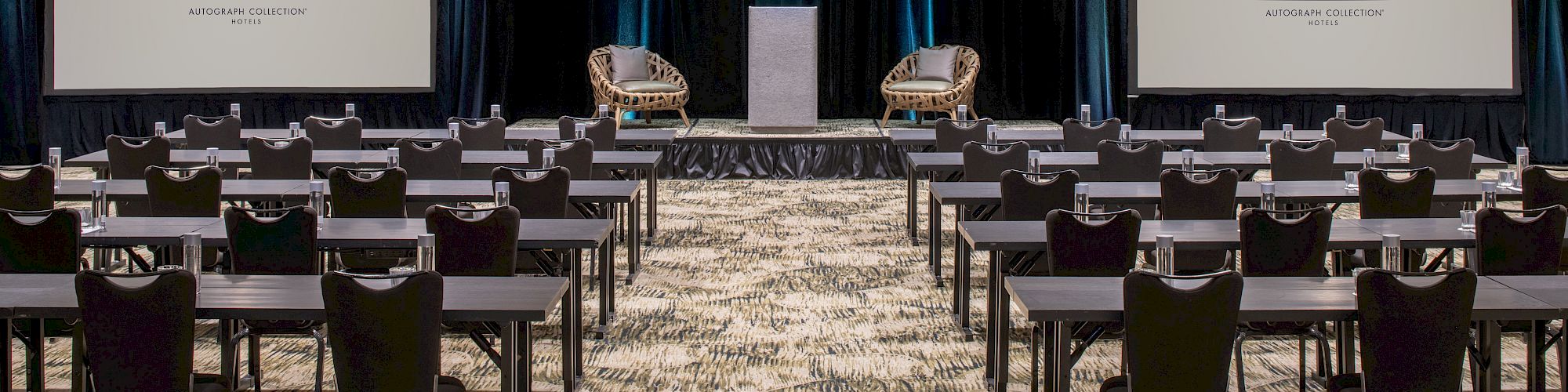 A conference room setup with rows of tables and chairs facing a stage with two screens labeled 