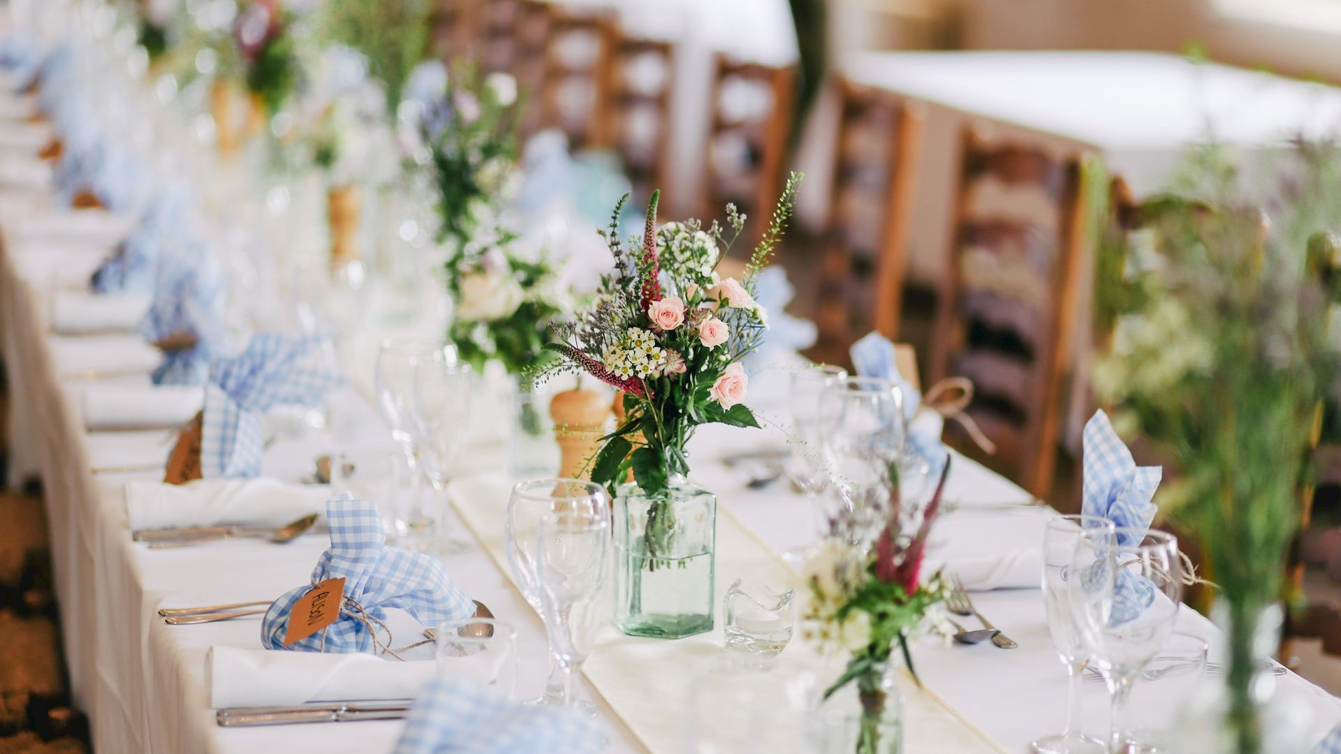 A beautifully set long dining table with white tablecloth, blue napkins, elegant glassware, and floral centerpieces in green vases, ready for a formal event.