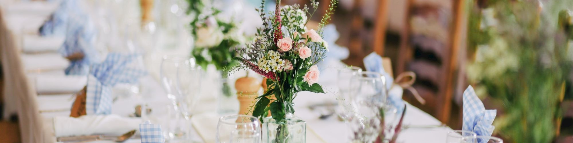 A beautifully set long dining table with white tablecloth, blue napkins, elegant glassware, and floral centerpieces in green vases, ready for a formal event.