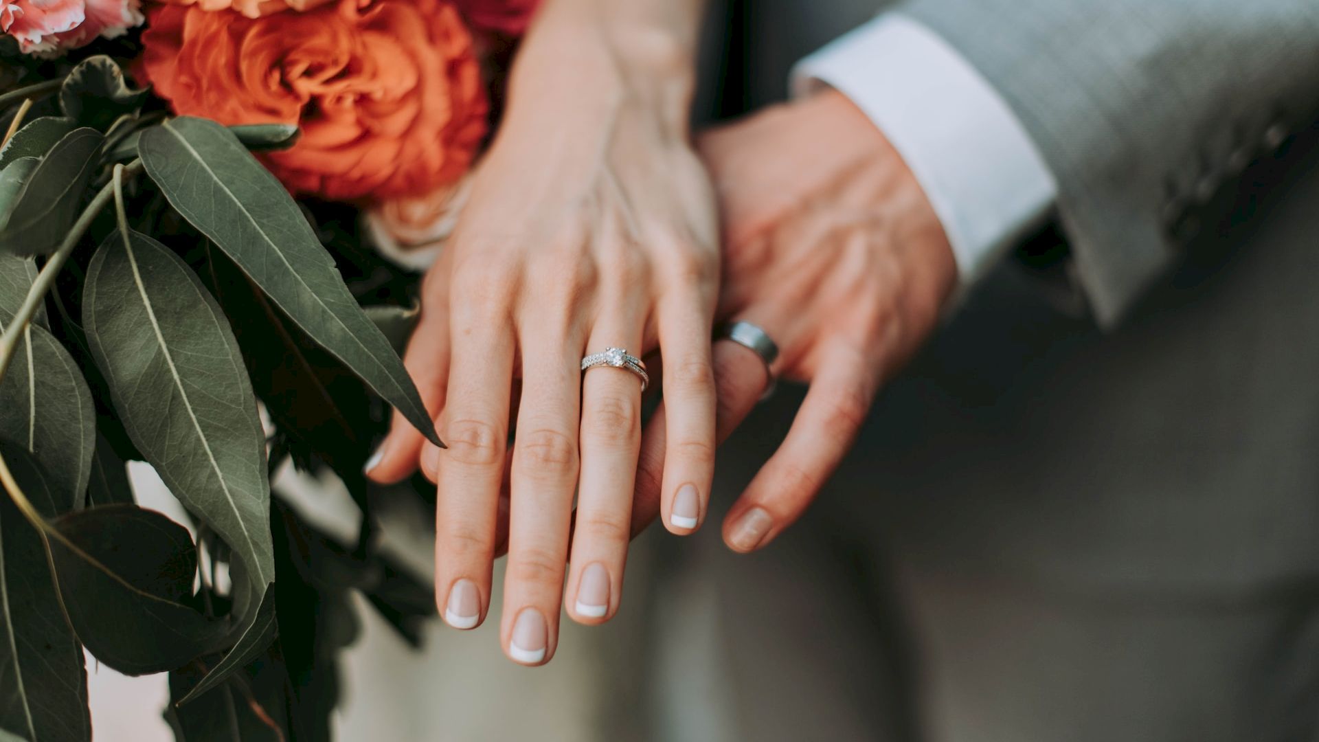 A close-up of two hands showing wedding rings, with a bouquet of flowers featuring red and orange roses in the background.