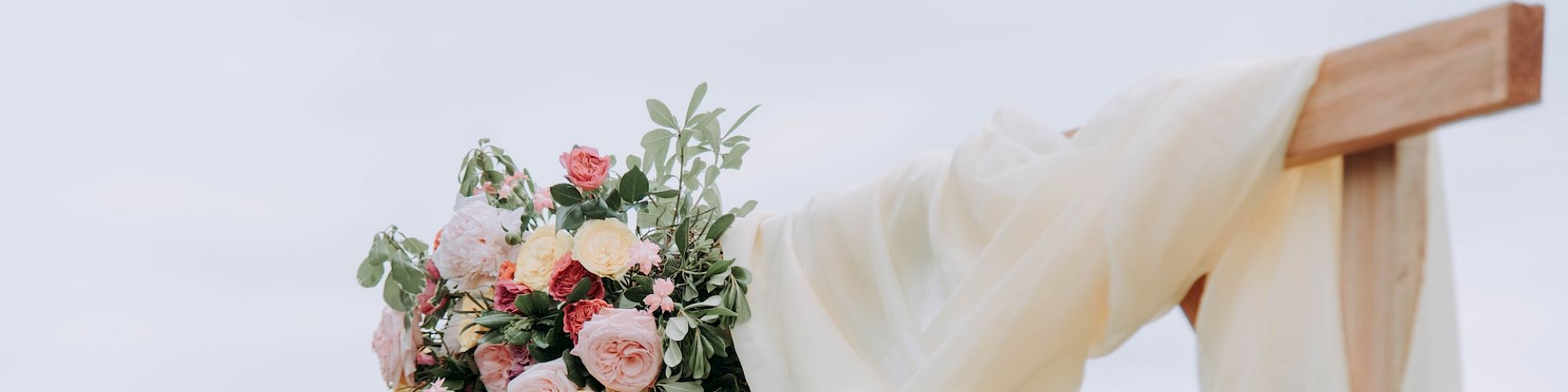 The image shows a wooden arch decorated with draped white fabric and a large bouquet of pink, white, and green flowers against a backdrop of greenery.