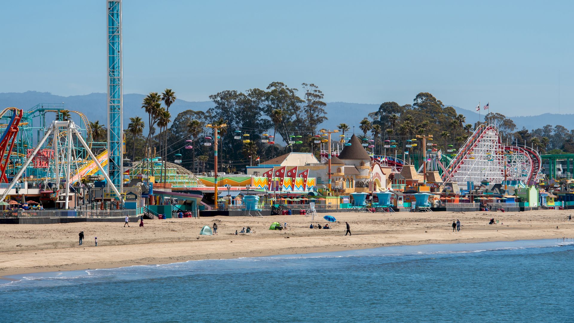 This image shows a beach with an amusement park featuring rides and attractions. People can be seen strolling along the shore and in the park.