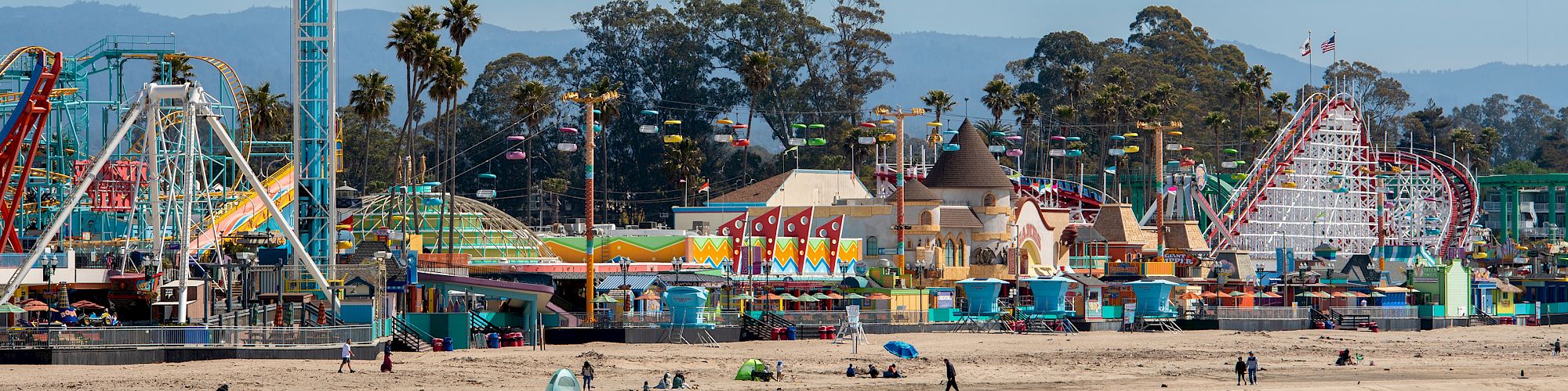 This image shows a beach with an amusement park featuring rides and attractions. People can be seen strolling along the shore and in the park.