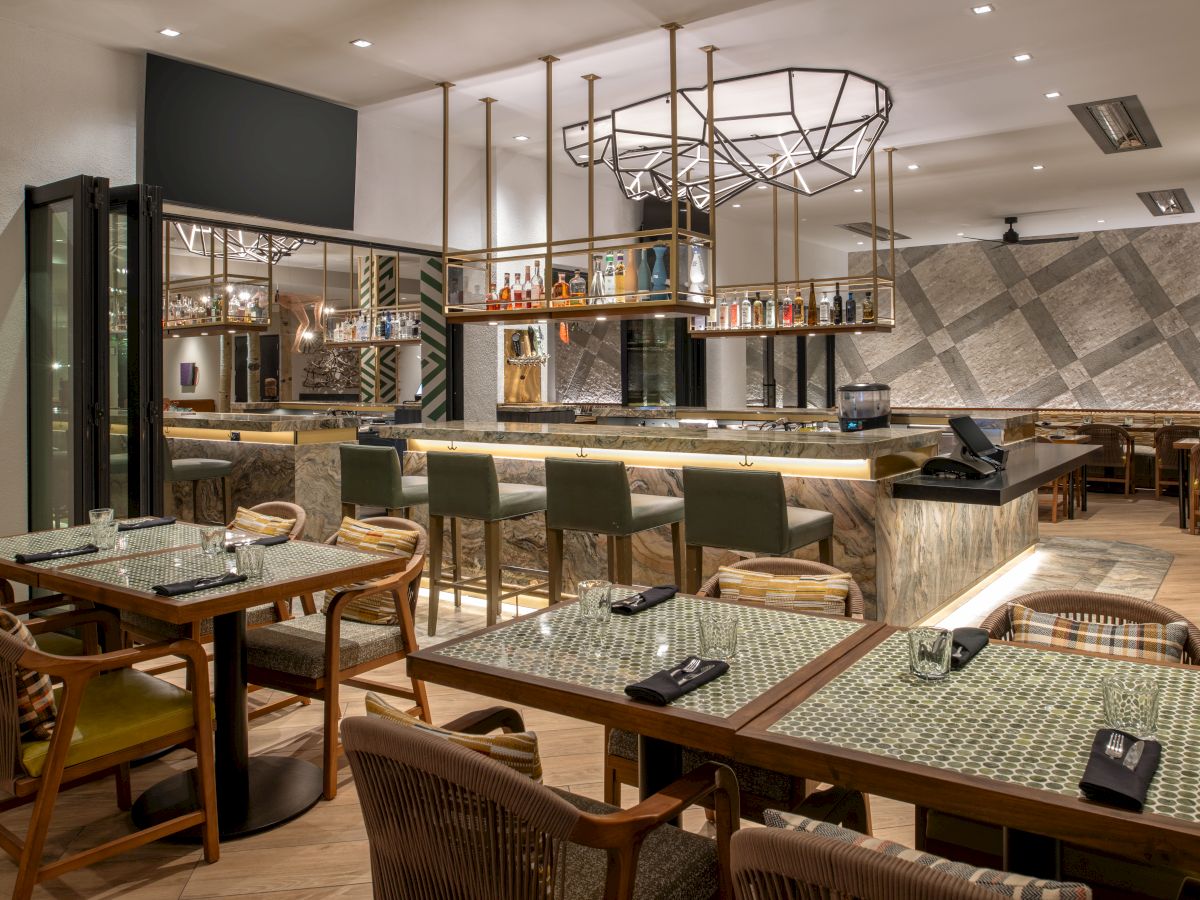 This image shows a modern, stylish restaurant interior featuring a bar area with high chairs, tables with woven chairs, and contemporary lighting.