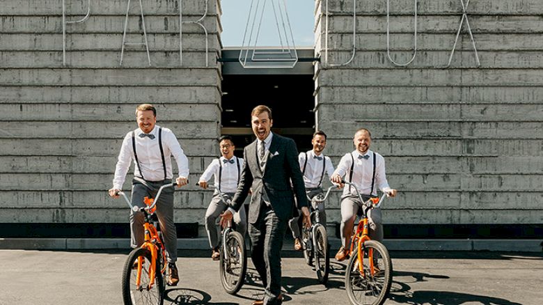 A man in a suit on a skateboard is flanked by four men on bikes, posing in front of a building with the word 