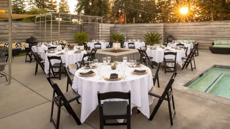 An outdoor dining setup features round tables with white tablecloths, black chairs, elegantly set dining ware, and a setting sun in the background.