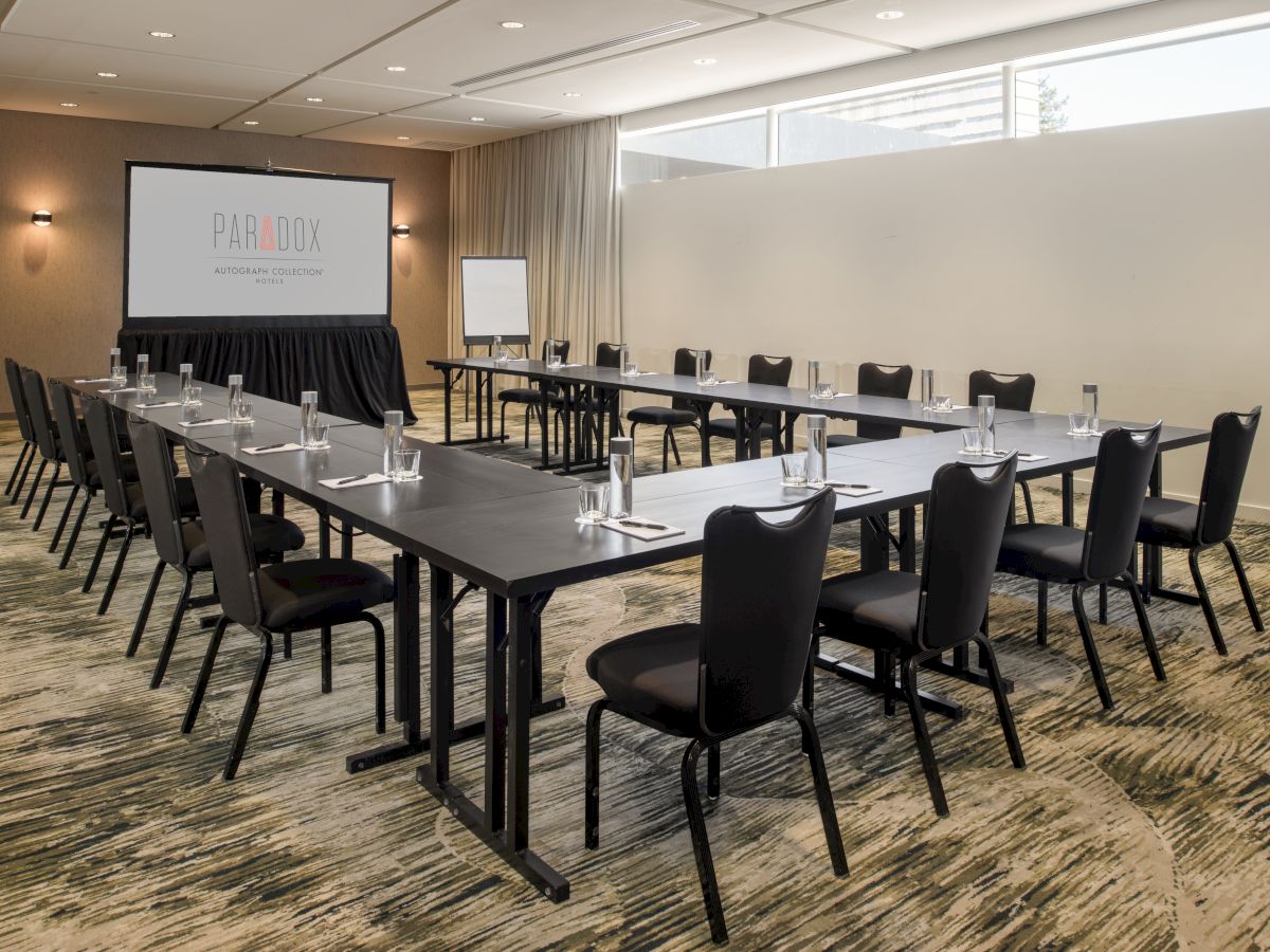 A conference room with U-shaped table setup, empty chairs, water glasses, a screen with "Paradox" logo, and a flip chart stand.
