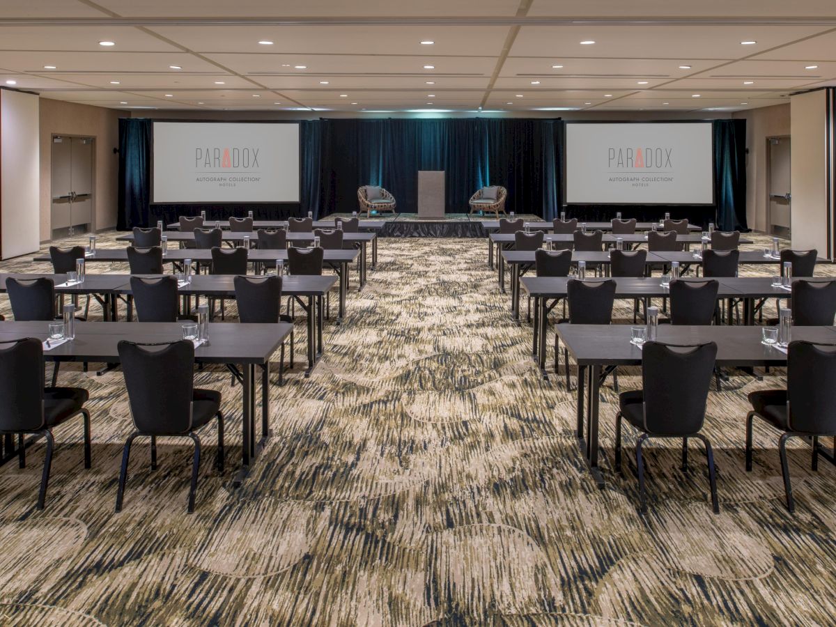 The image shows a conference room setup with chairs and tables facing a stage with two screens displaying "Paradox" and a podium in the center.