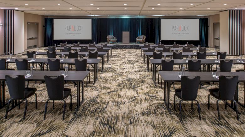 The image shows a conference room setup with chairs and tables facing a stage with two screens displaying 