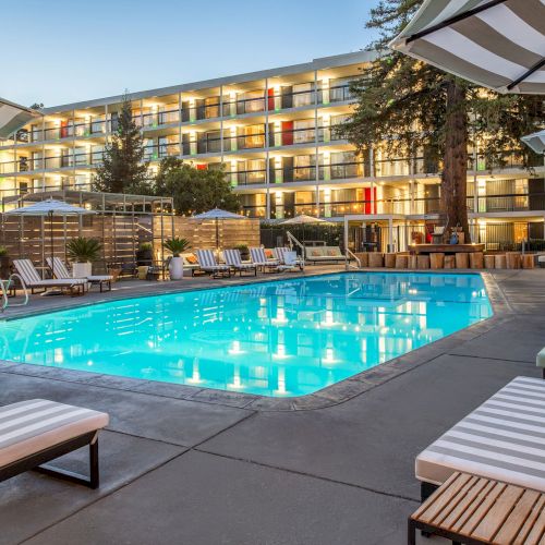 A luxurious outdoor pool area surrounded by lounge chairs and umbrellas, adjacent to a modern hotel building with multiple balconies and evening lighting.