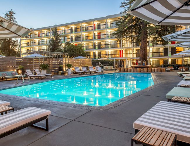 A luxurious outdoor pool area surrounded by lounge chairs and umbrellas, adjacent to a modern hotel building with multiple balconies and evening lighting.