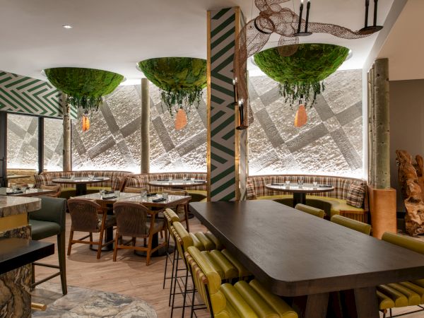 A modern restaurant interior with unique decor, green hanging plants, patterned walls, and a mix of seating options, including a long communal table.