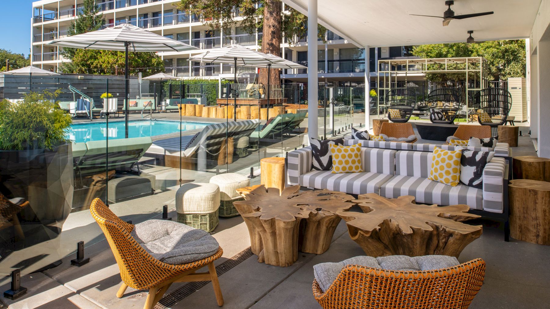 The image shows an outdoor seating area near a swimming pool, with wicker chairs, wooden tables, and a striped couch under a shaded patio area.