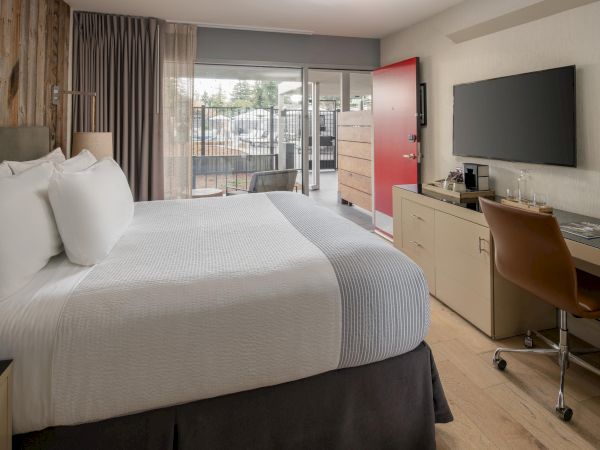 A modern hotel room features a large bed, TV, desk, and chair, with a red door in the background, leading to an outdoor area with a view.