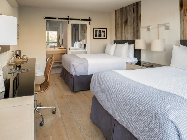 The image shows a hotel room with two double beds, a desk with a chair, a mirror, lamps, and modern decor. The room is bright and tidy.