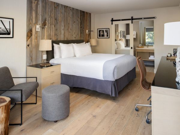 The image shows a modern hotel room with a bed, a desk, angled chair, a wooden stool, and framed artwork on the walls, ending the sentence.