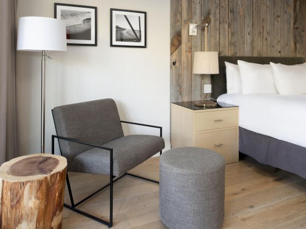 The image shows a modern hotel room with a bed, chair, side table, lamp, and wooden decor. There are two framed pictures on the wall.