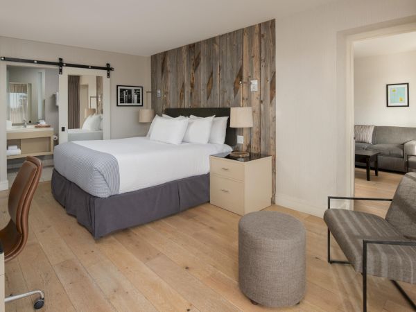 A modern hotel room with a bed, desk, and chair, featuring wooden accents and a sliding door leading to a bathroom and a seating area.