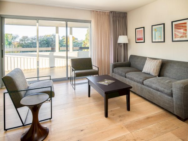 A modern living room with a grey sofa, two grey armchairs, a wooden coffee table, a side table, and framed art on the wall, overlooking a balcony.