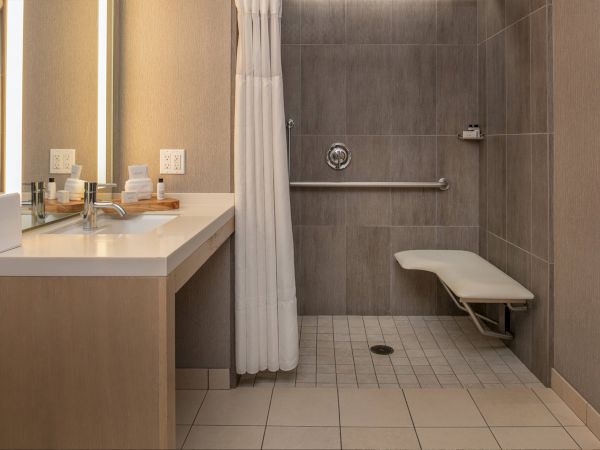 This image shows an accessible bathroom with a roll-in shower, a wall-mounted shower bench, grab bars, a sink with countertop space, mirror, and toiletries.