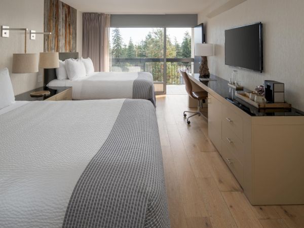 A modern hotel room with two beds, a flat-screen TV, a desk, and a floor-to-ceiling window with a view of the trees outside.
