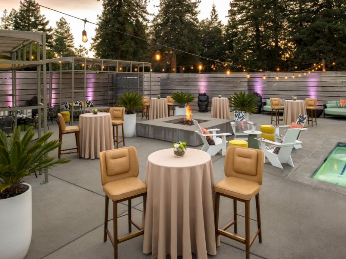 Outdoor patio with high tables and chairs, a fire pit, string lights, potted plants, and a small pool, ideal for social gatherings or events.