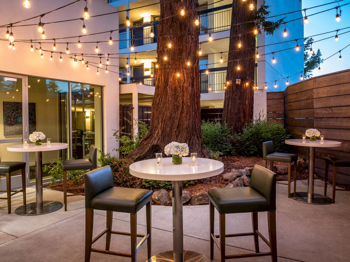 An outdoor patio with string lights features round tables, chairs, and small floral arrangements. Trees and building windows are visible in the background.