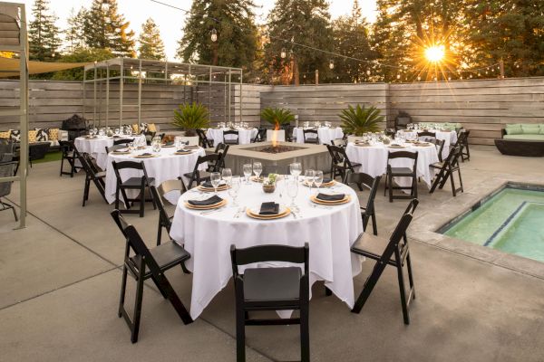 An outdoor event setup with round tables, black chairs, white tablecloths, a fire pit, and string lights in a patio area surrounded by trees.