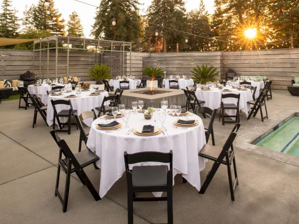 An outdoor event setup with round tables, black chairs, white tablecloths, a fire pit, and string lights in a patio area surrounded by trees.